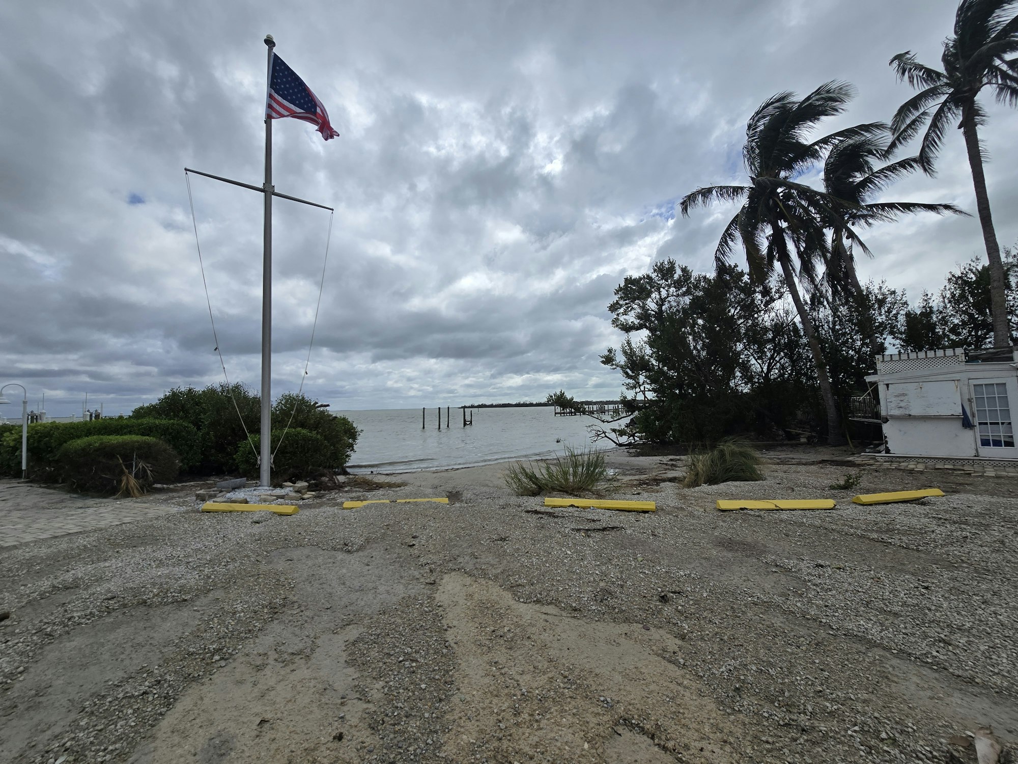 A cloudy coastal scene with an American flag, palm trees, a small building, and a rocky shoreline.