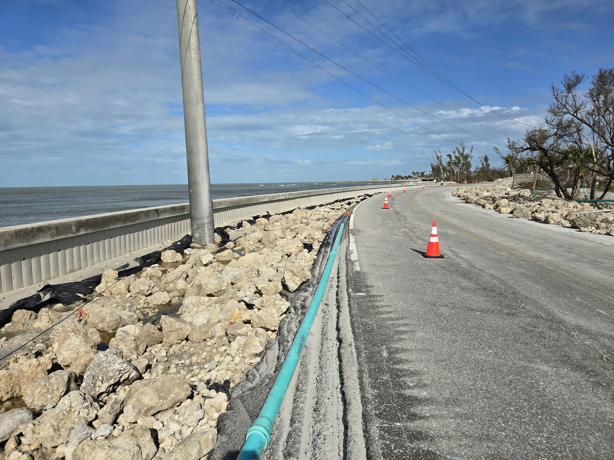 Coastal road with traffic cones, rocky shoreline, power lines, and blue sky in the background.