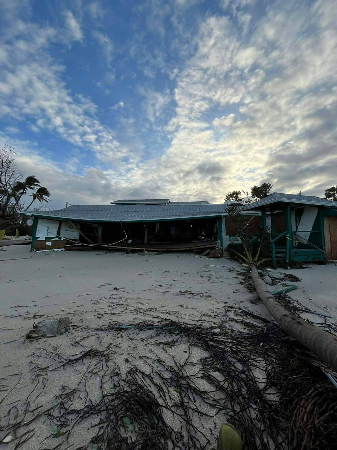 A damaged beachfront building with debris and fallen palm trees, under a cloudy sky.