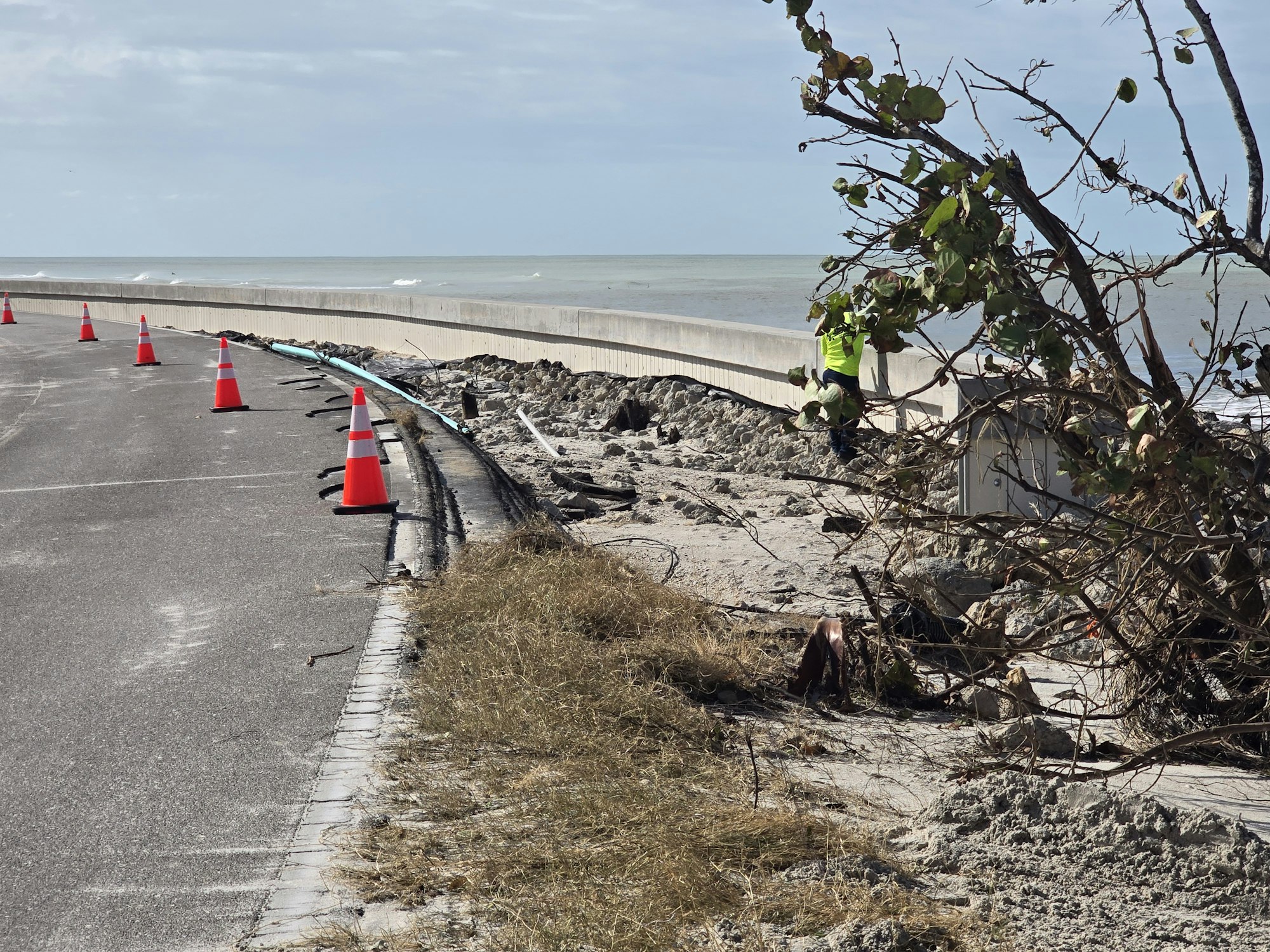 Roadside with orange traffic cones, erosion damage, coastline in the background, and a worker near a damaged area.