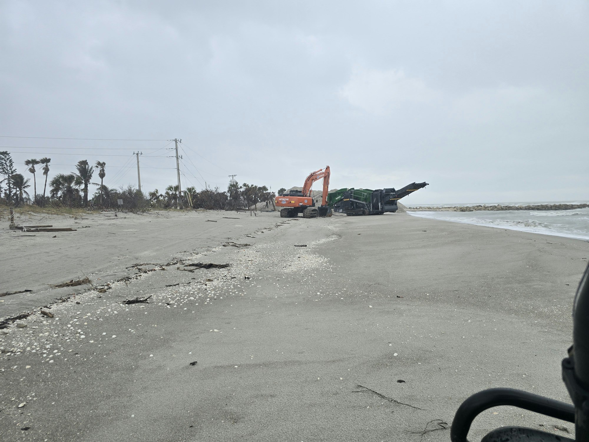 A beach with a construction vehicle and debris, overcast sky, and palm trees in the background.