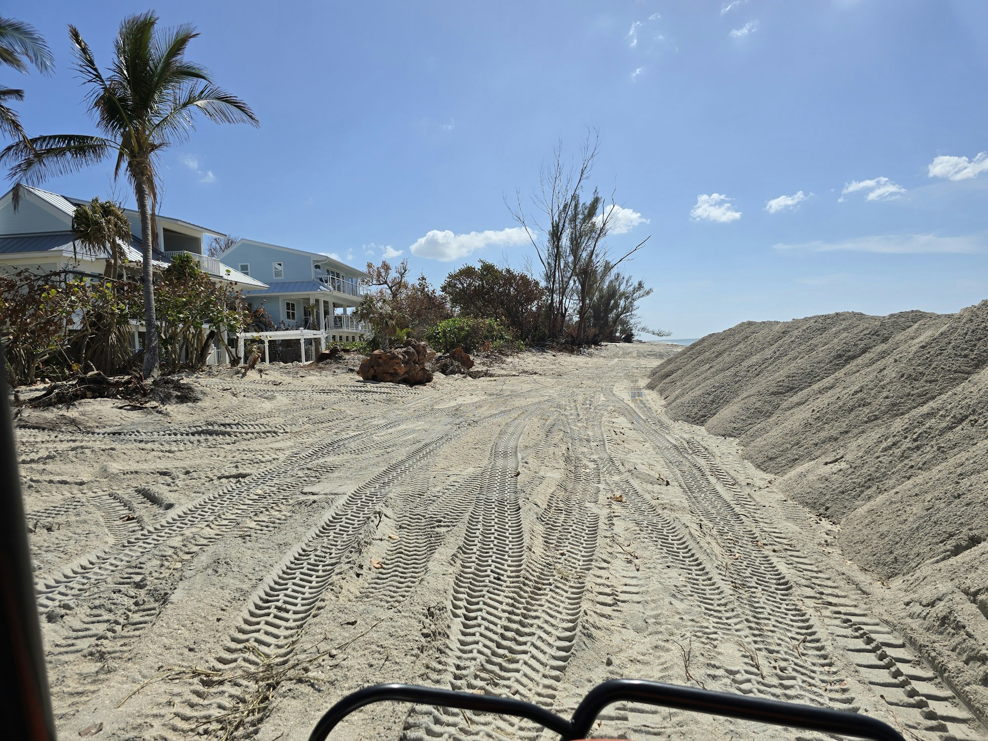 Beachfront homes, palm trees, and vehicle tire tracks on sandy terrain with mounds of sand under a blue sky.