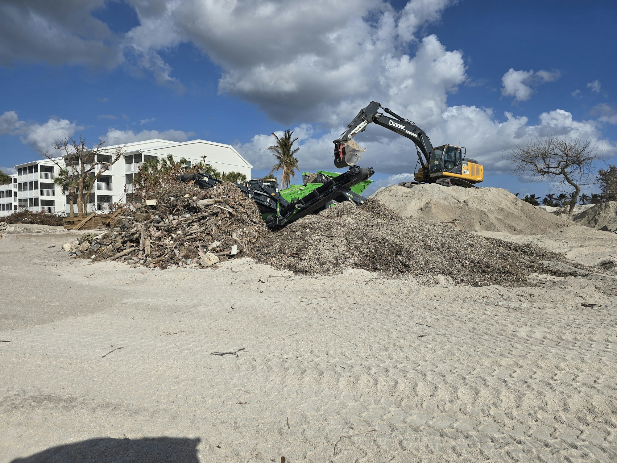 An excavator moves debris on a sandy beach near buildings and a tree under a cloudy sky.