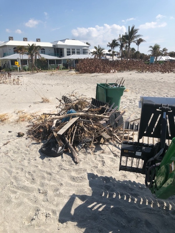 Beach debris near a house, including wood and trash, with a green garbage bin and a tractor attachment.