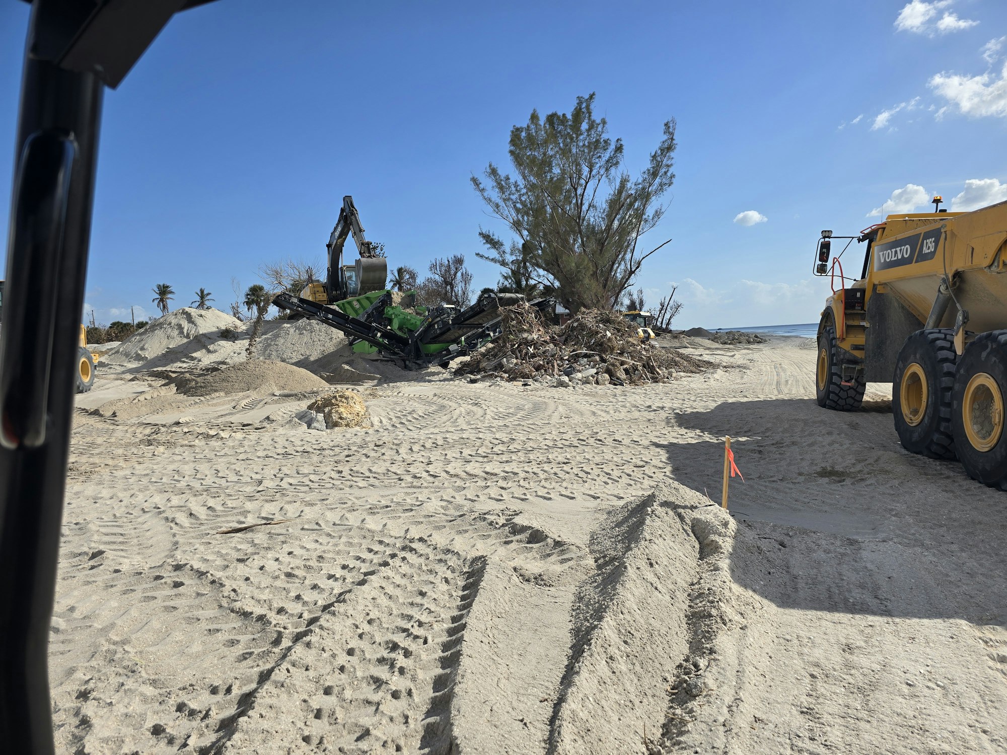 Construction site with heavy machinery, sand piles, and tire tracks under a clear sky.