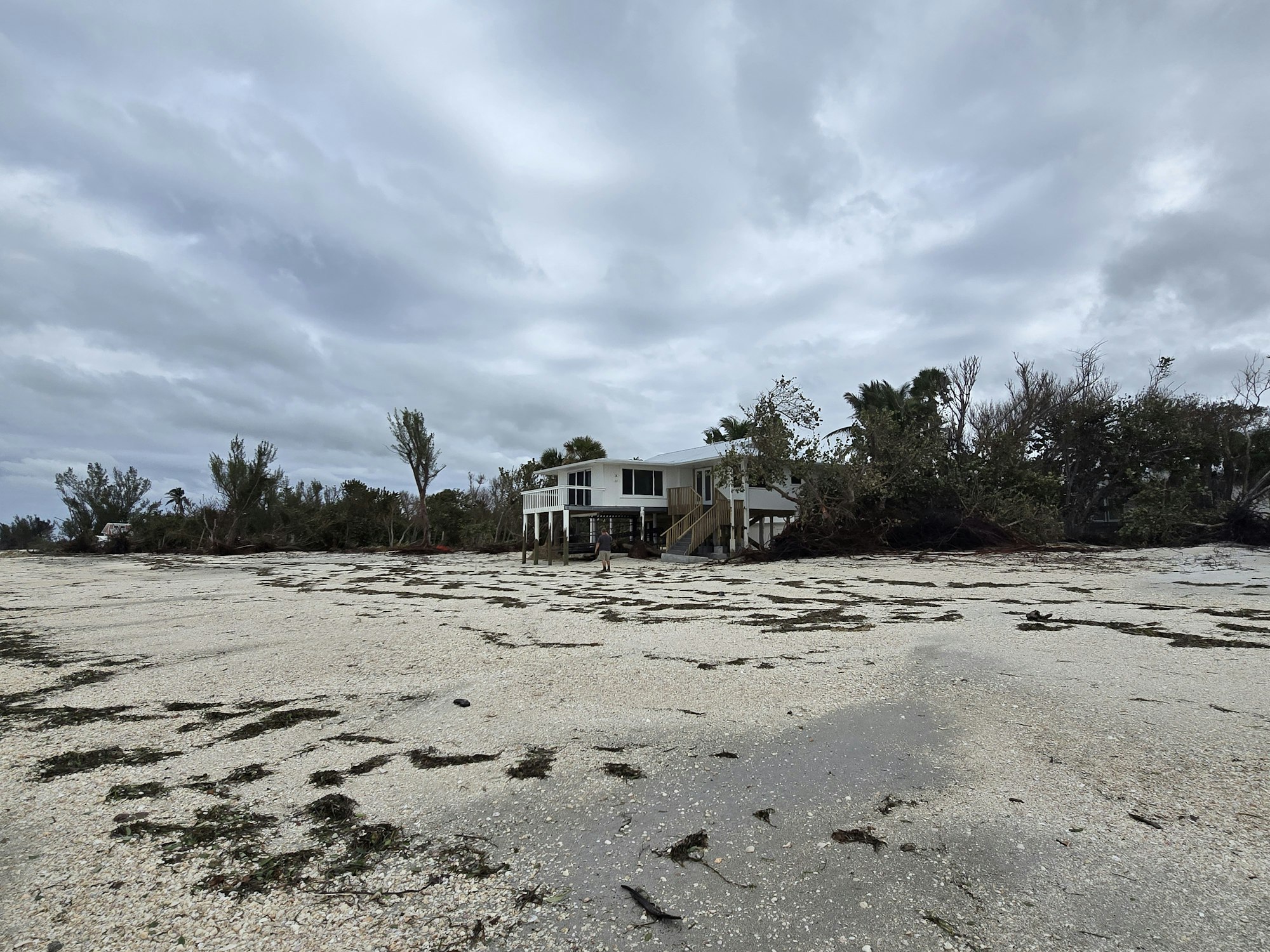A house on stilts by a sandy area with scattered debris and overcast sky.