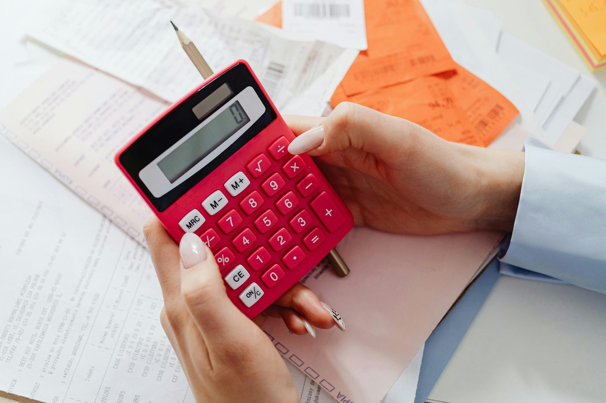A person holds a pink calculator and a pencil over a pile of receipts and papers, likely for budgeting or calculations.
