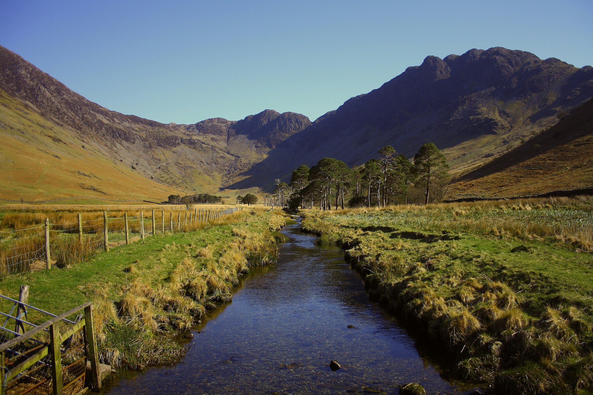 A serene landscape featuring mountains, a meandering stream, grassy fields, and trees under a clear blue sky.