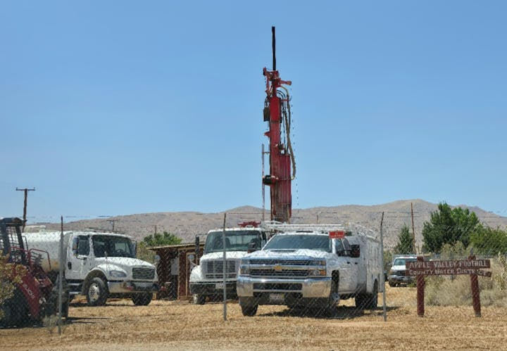 White trucks and a red drilling rig in a fenced area labeled "Apple Valley Foothill County Water District" against a mountainous backdrop.