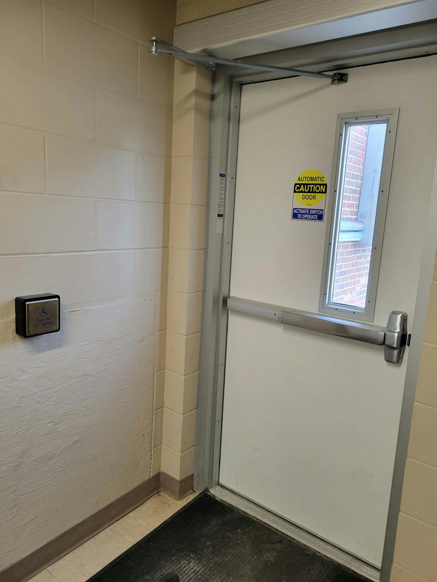 The image shows a hallway with an automatic door, a caution sign, and a button for accessible entry.