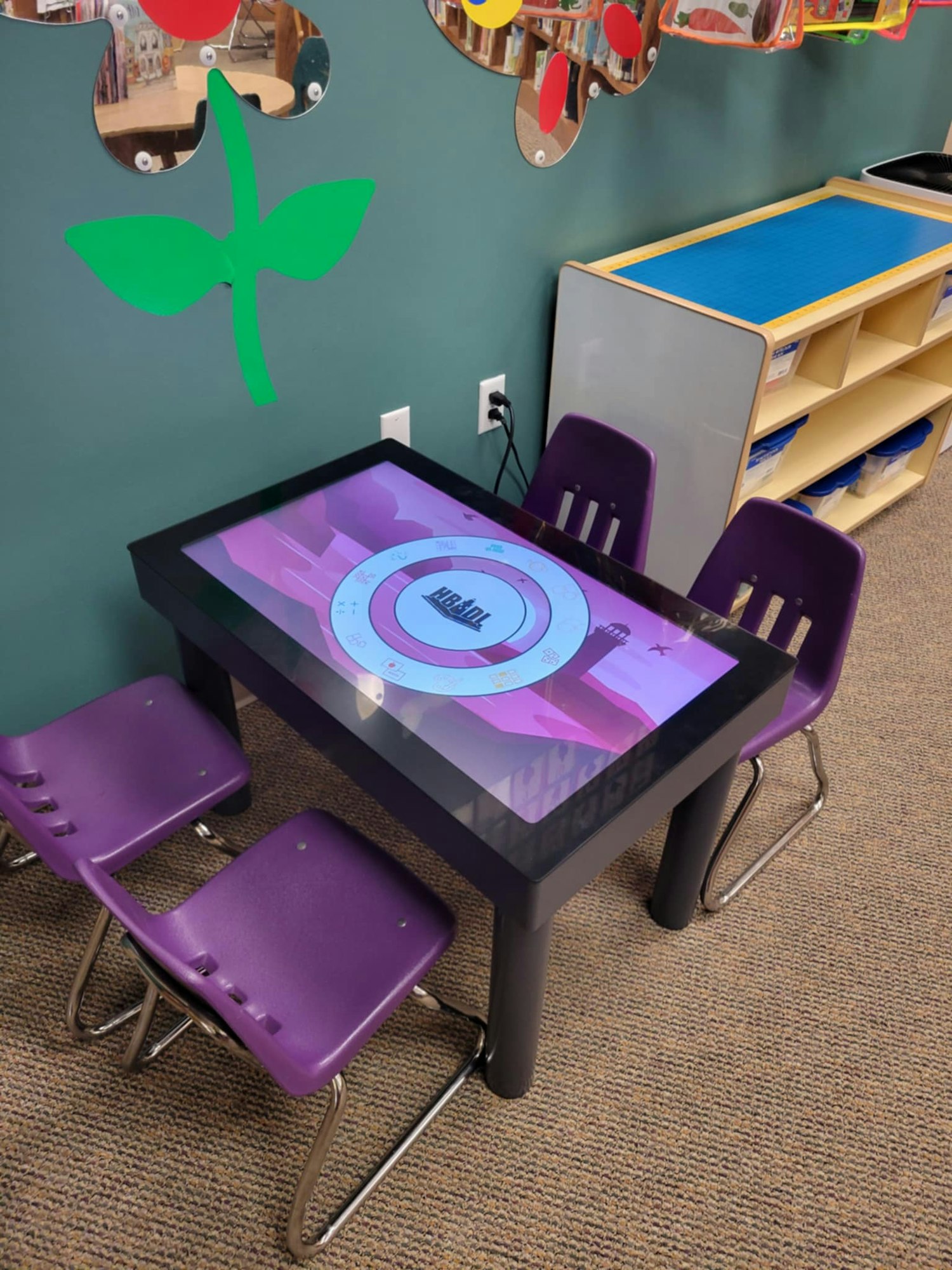 A touchscreen table with interactive content, surrounded by four purple chairs, in a children's activity area.