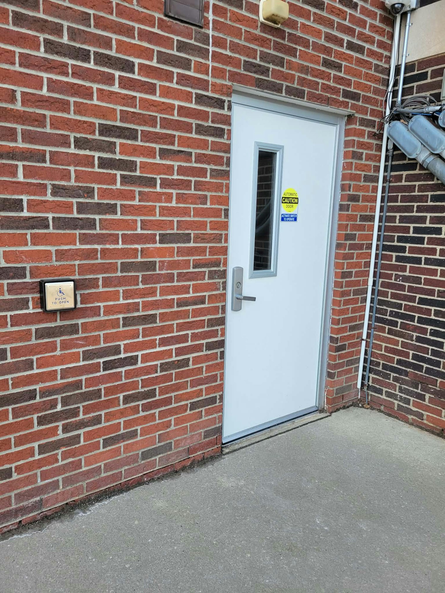 A white door in a brick wall, featuring a caution sign and a push button next to it.