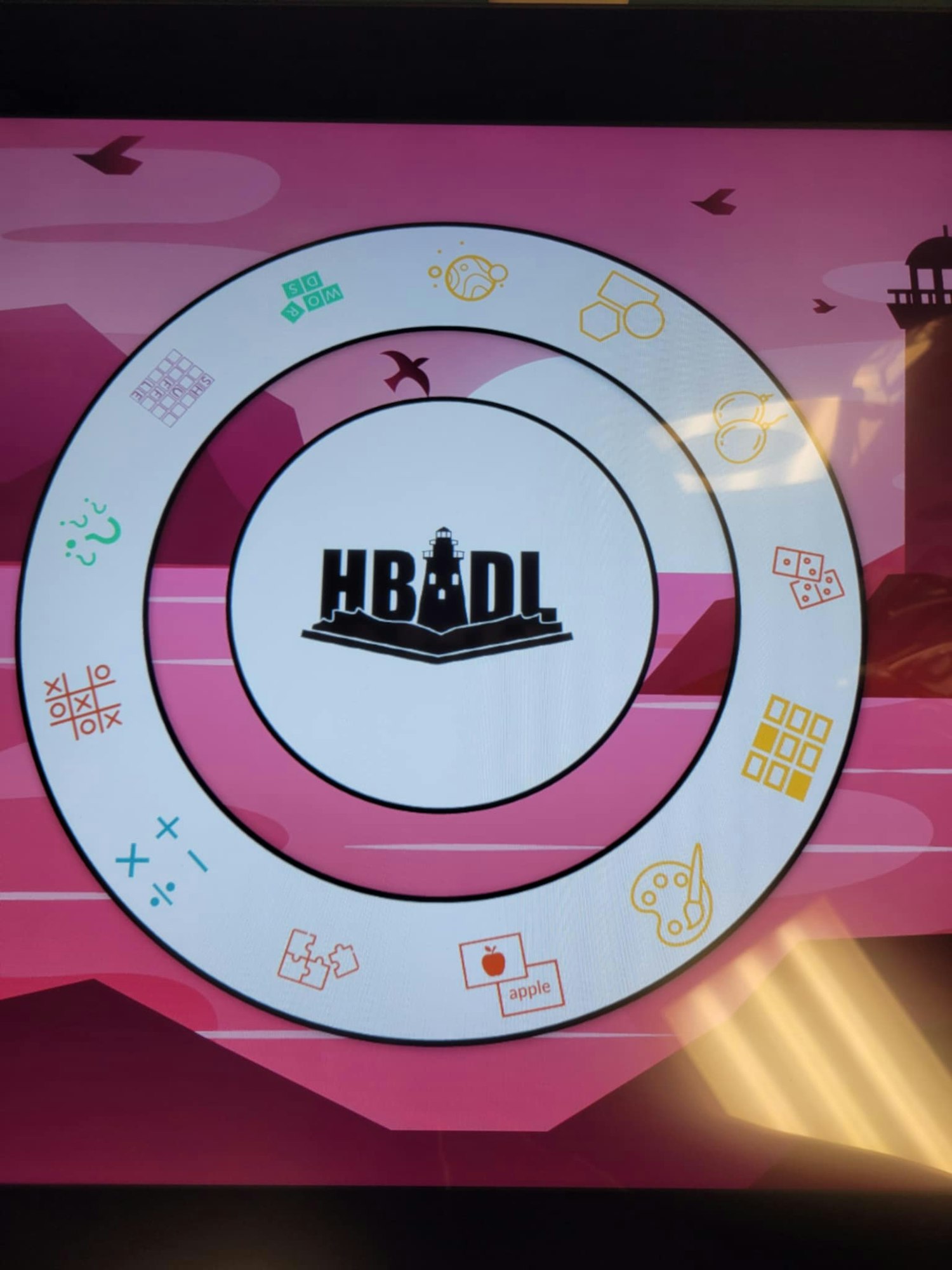 A circular game interface with various icons (sports, puzzles, math) and "HBADL" in the center, set on a pink background with birds.