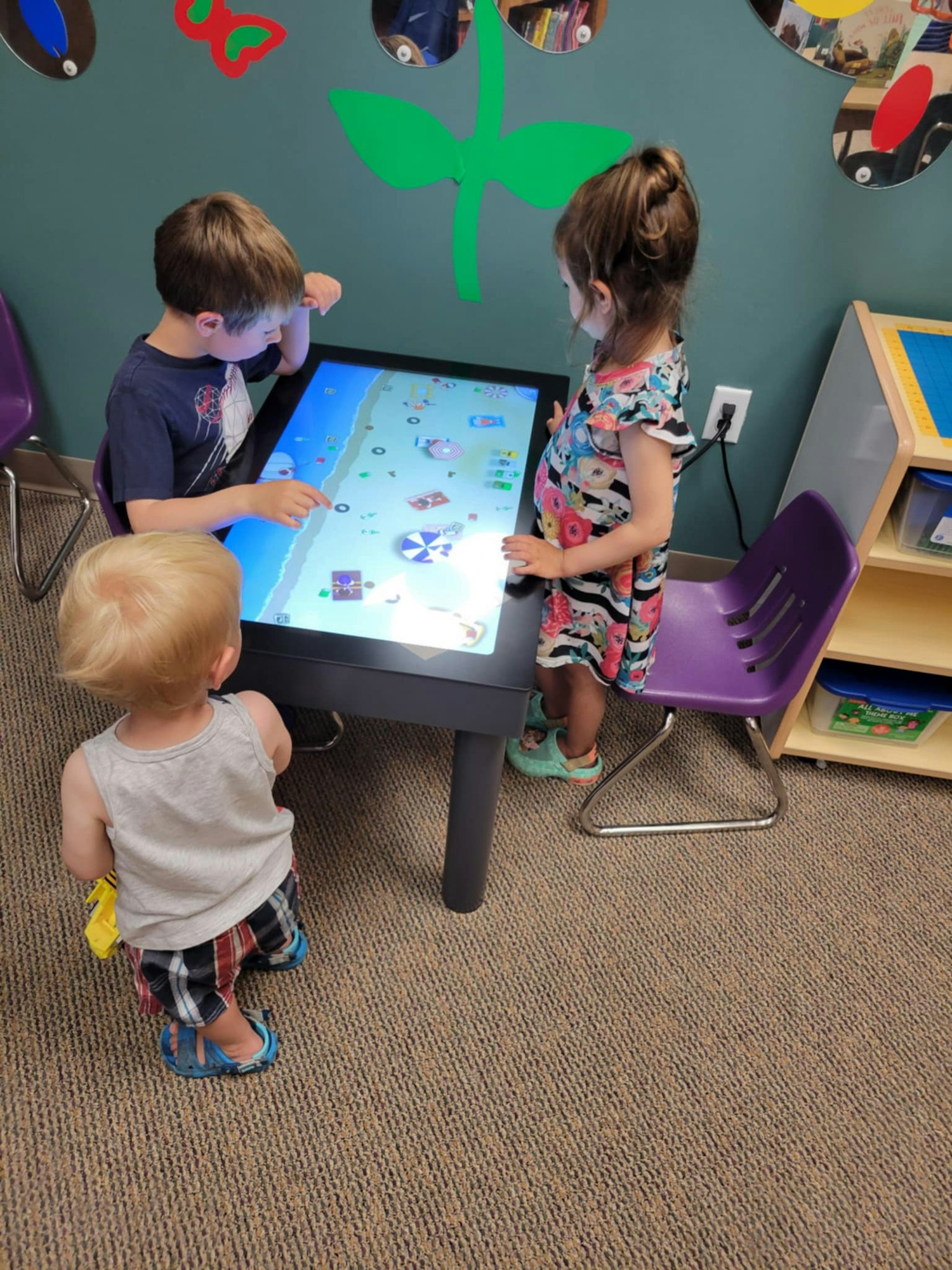 Three children engaged with an interactive table in a playroom, decorated with wall designs and mirrors.