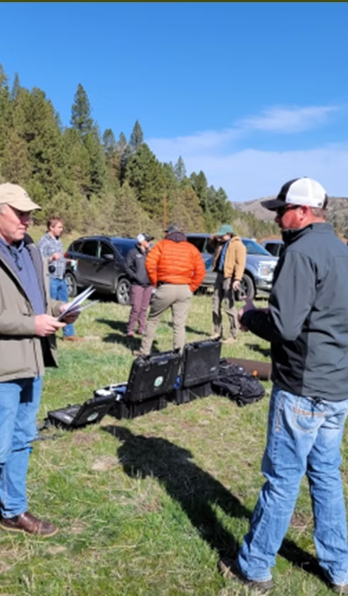 Group of people outdoors in a grassy area with equipment cases and parked vehicles. Trees and clear sky in the background.