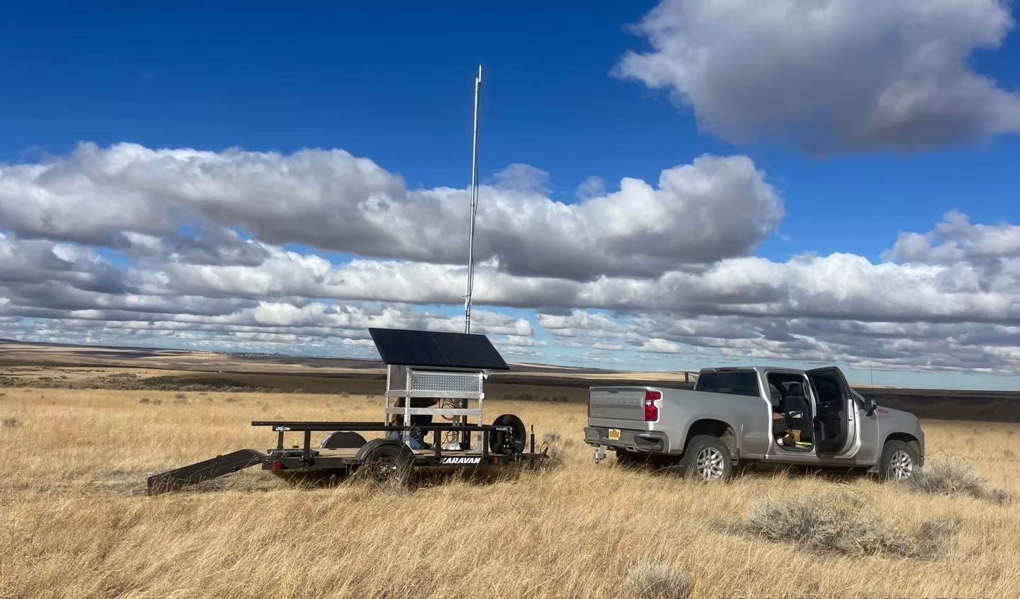 A pickup truck and a trailer with a solar panel and antenna on a grassy field under a cloudy sky.