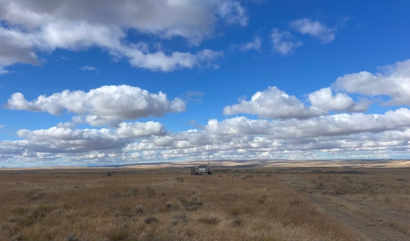 A vast, open field under a blue sky filled with fluffy clouds, with a structure and antenna in the distance.