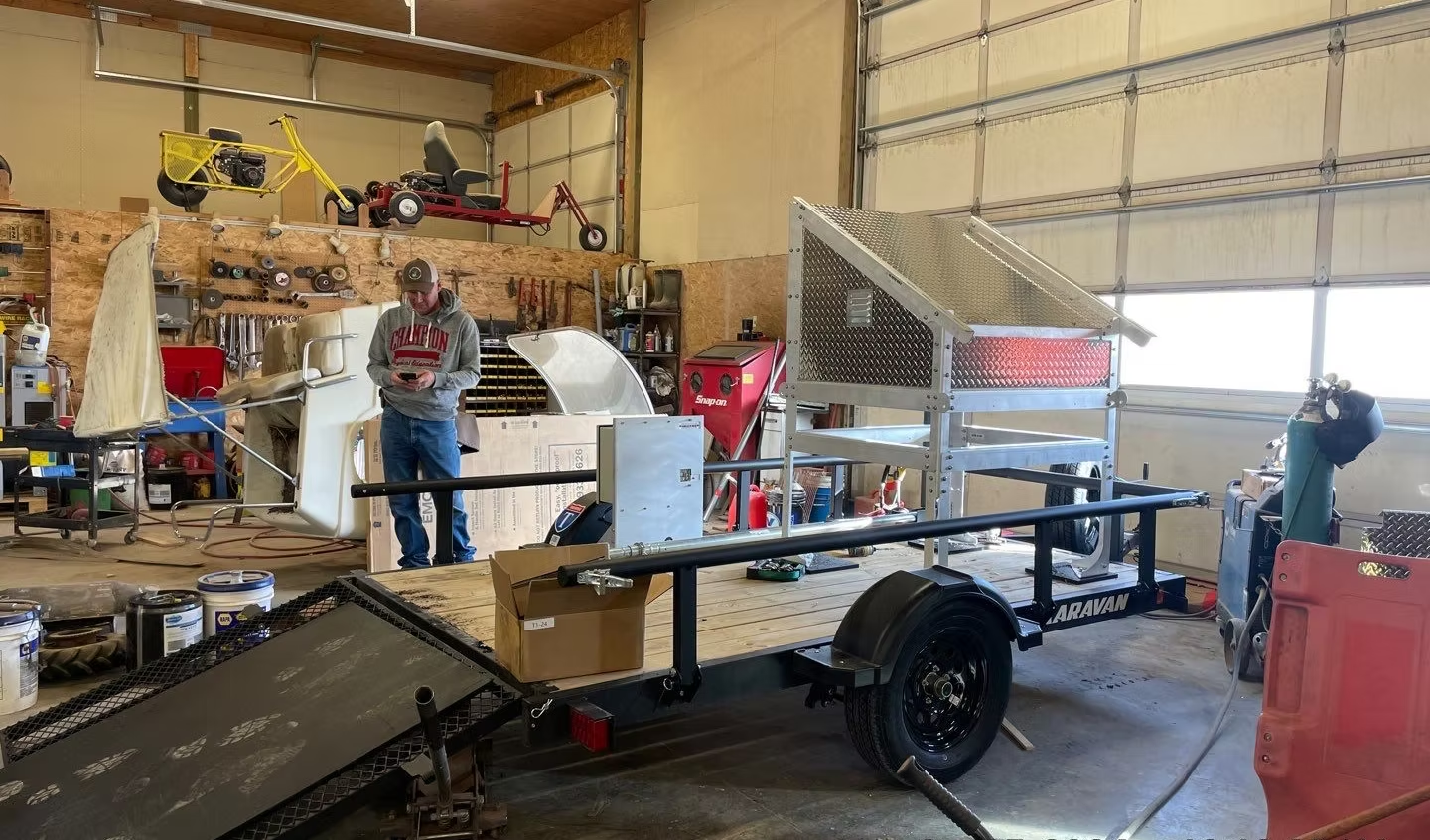 A person reads in a garage with tools, a trailer, bikes on a shelf, and other equipment around.