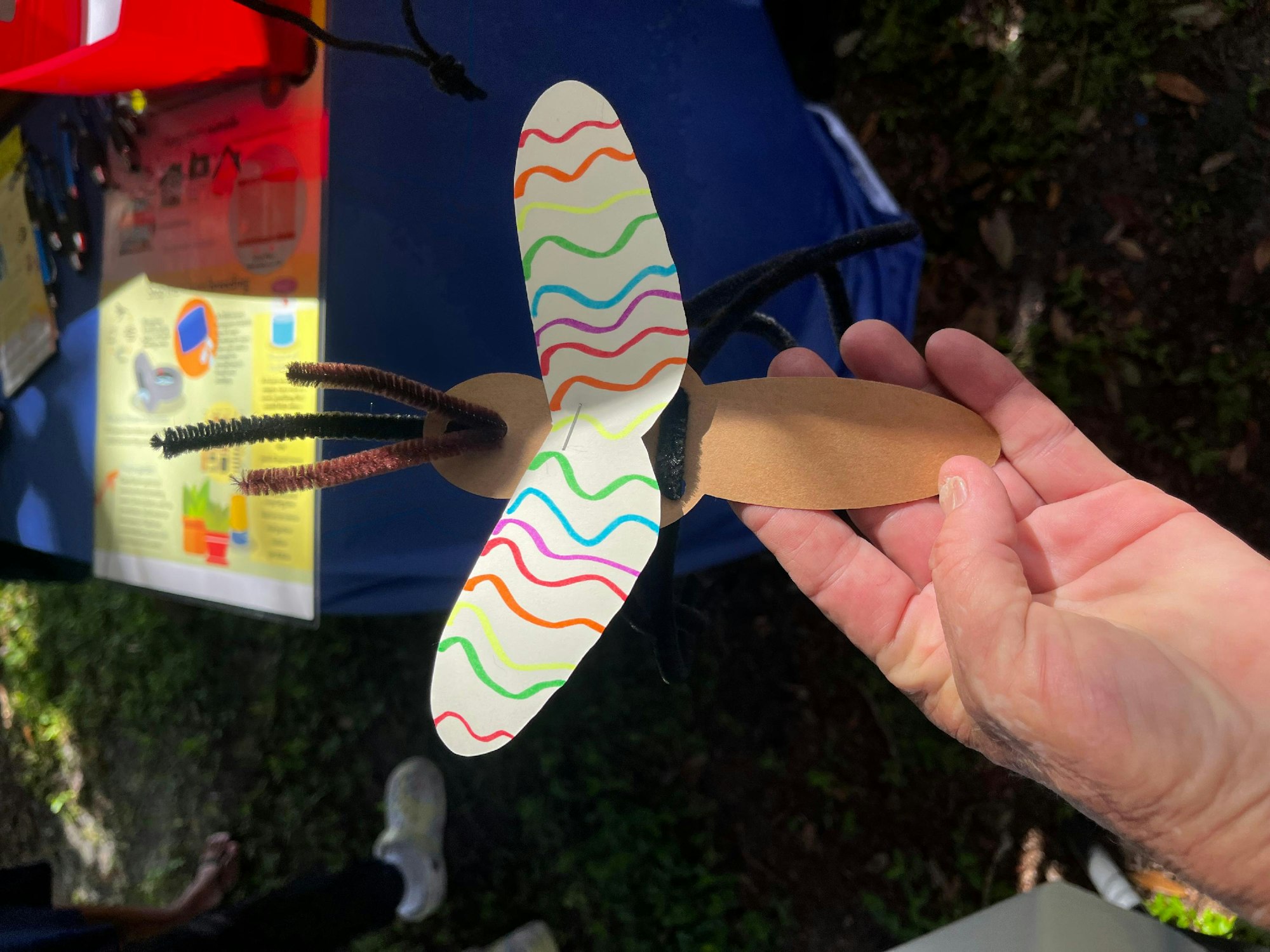 A hand holding a colorful, handmade paper dragonfly with pipe cleaner antennae.