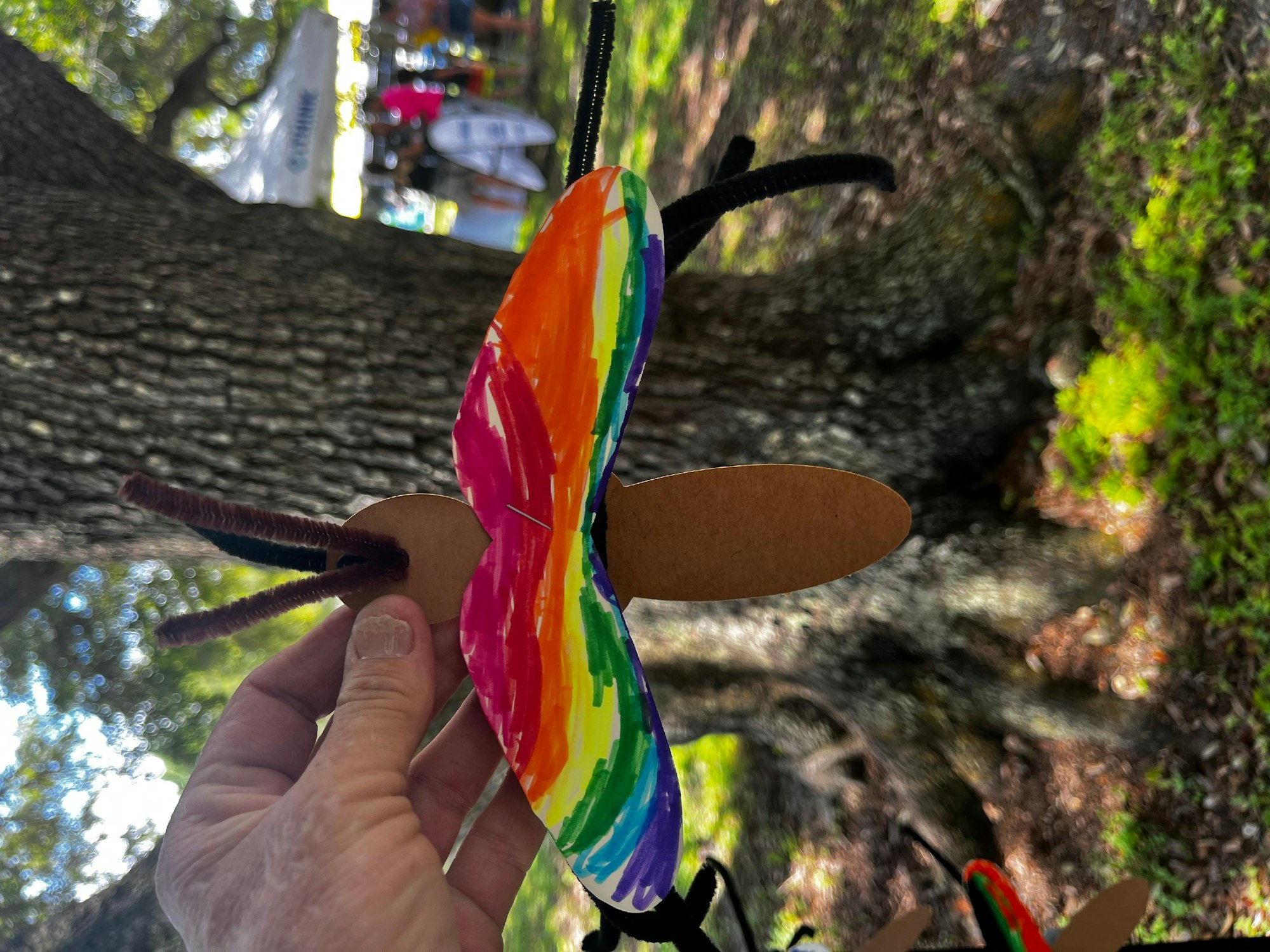 A hand holding a colorful, handmade butterfly craft with a tree and sunlight in the background.