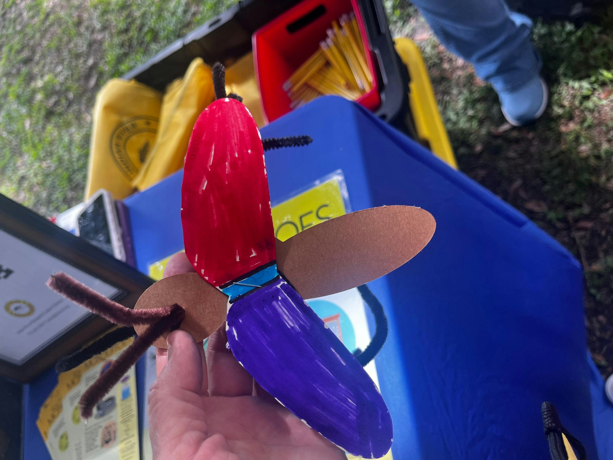 A hand holding a colorful, handmade rocket model against a blurred background of educational materials and a person's leg.