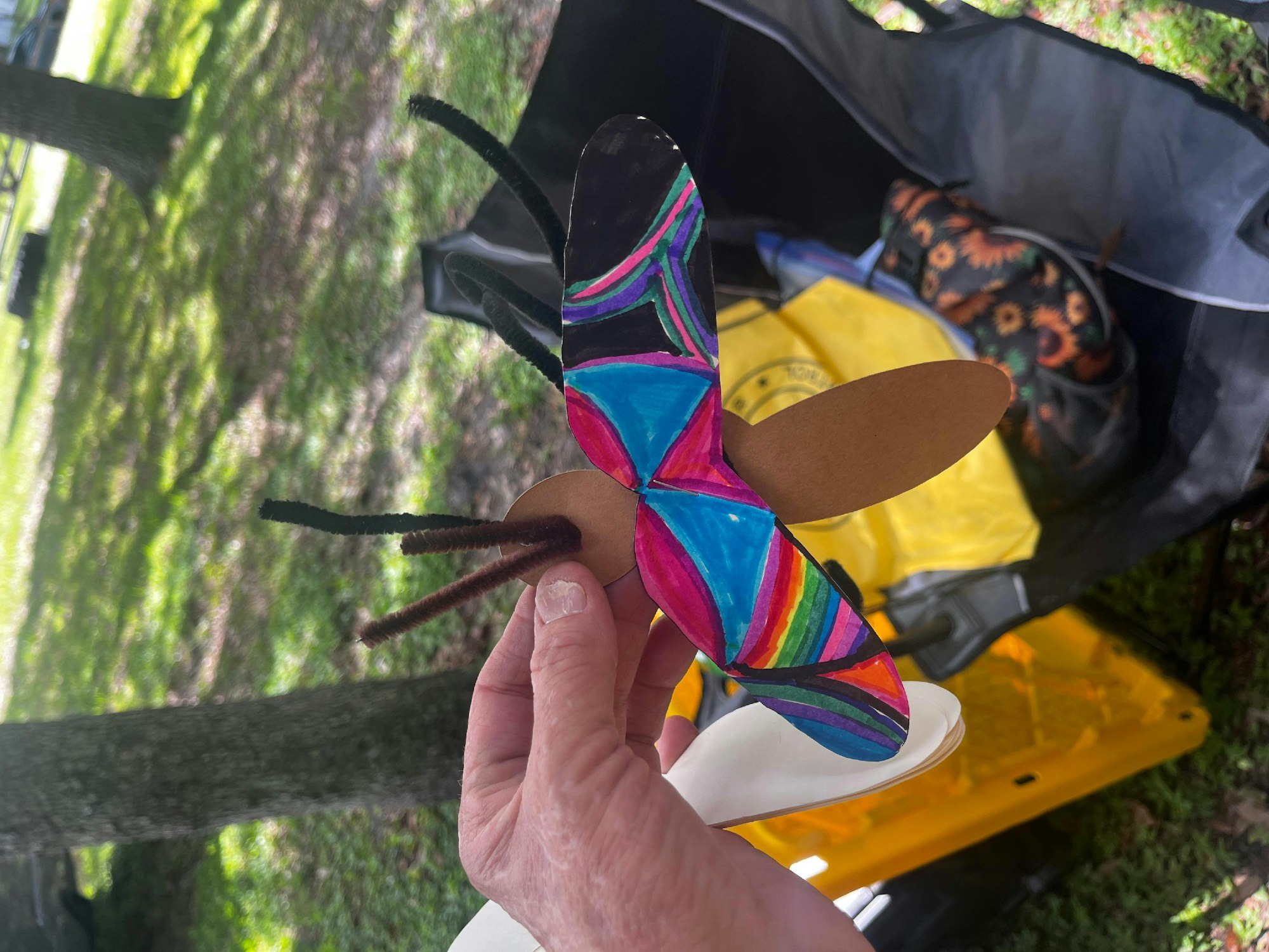 Colorful butterfly craft held by a person with a bag and yellow object in the background.
