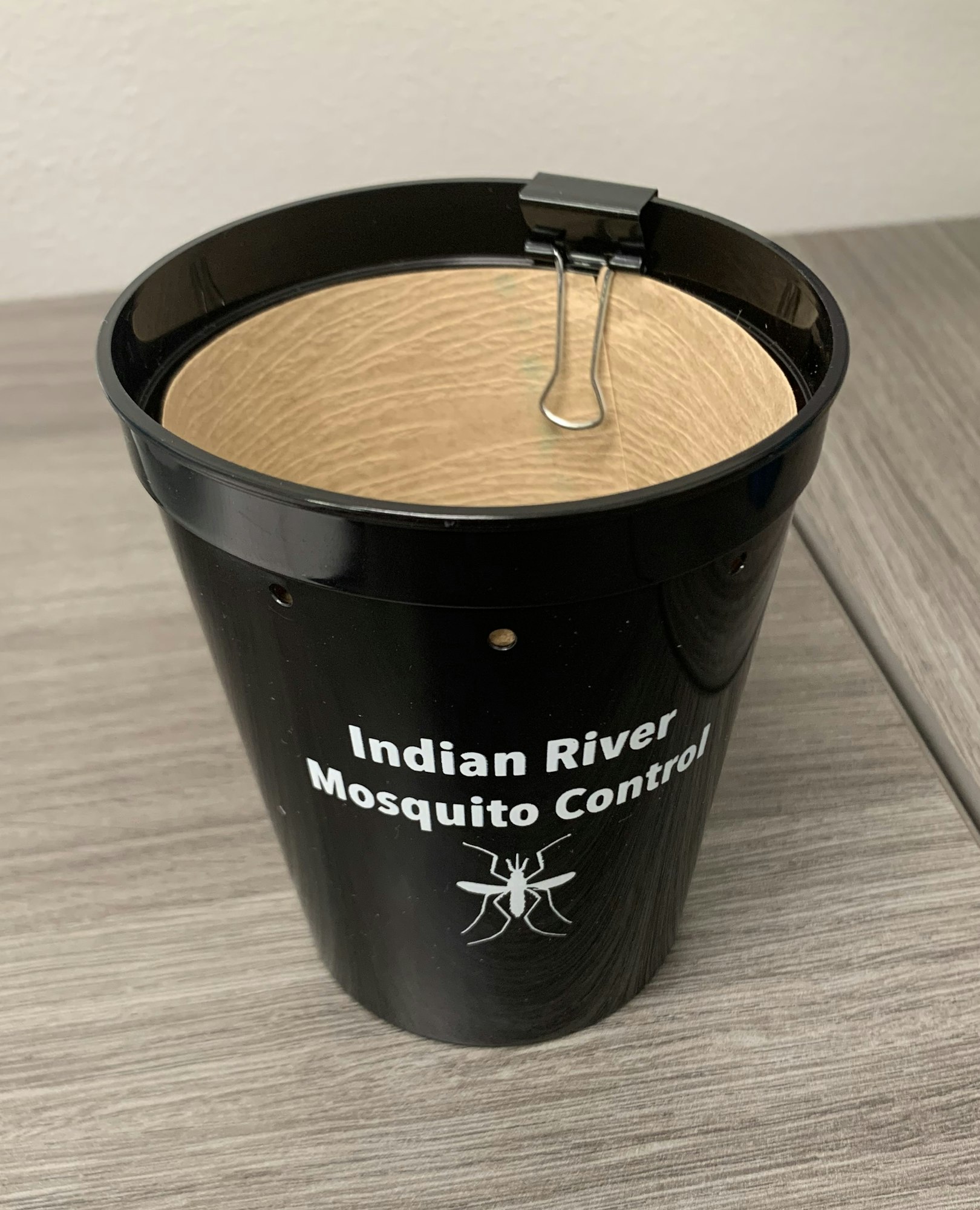 A black cup with "Indian River Mosquito Control" text and mosquito graphic, covered with a binder clip and tan mesh.
