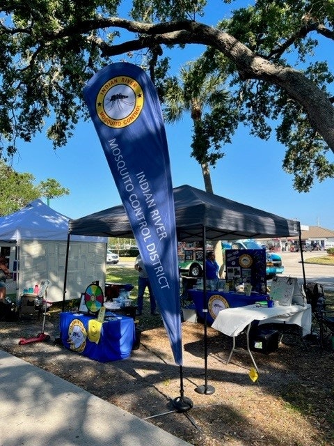 An outdoor event booth with a tall banner reading "Indian River Mosquito Control District," under a shaded canopy with tables and displays.