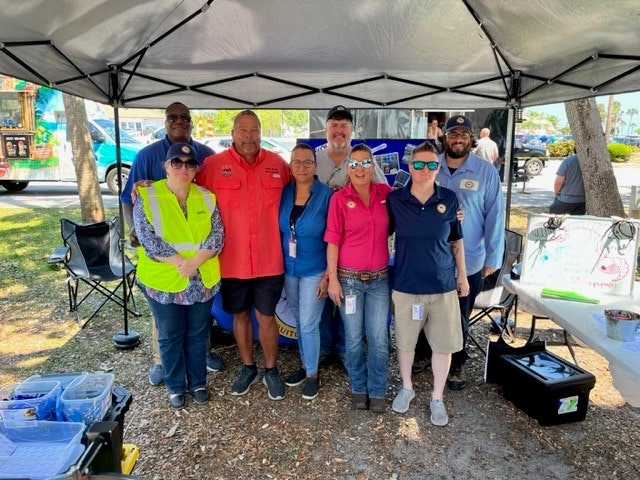 Group of people under a canopy, with a table and materials, possibly at an event or gathering.