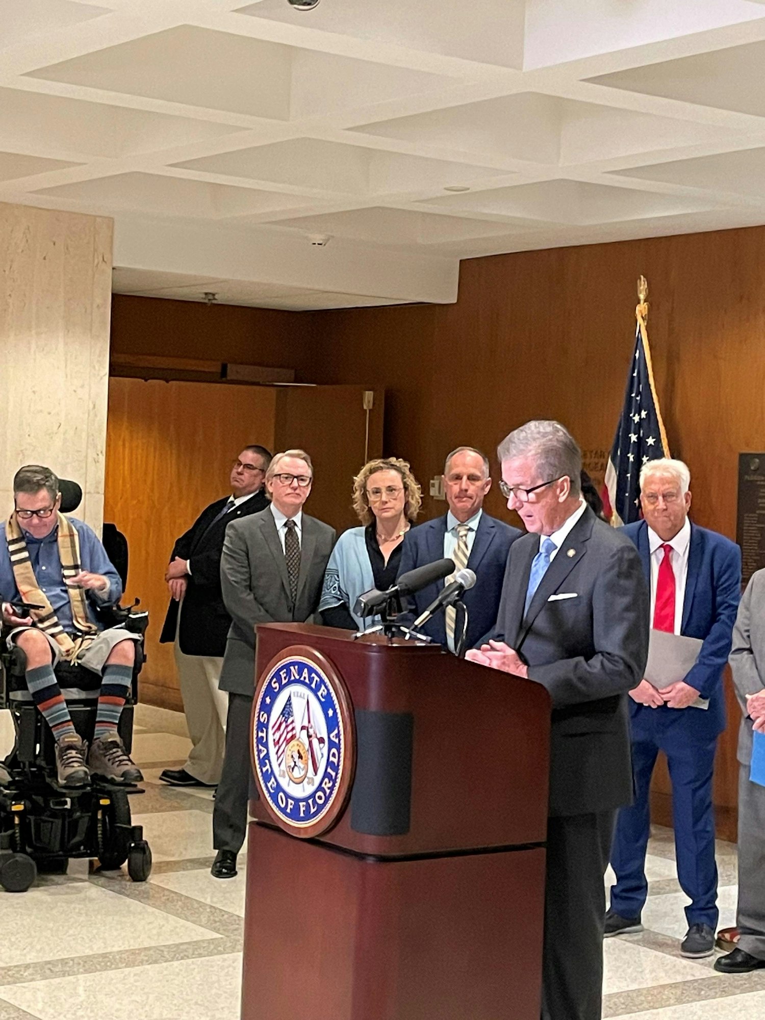 A man is speaking at a podium with the Florida Senate seal, while other individuals stand behind him, and a US flag is visible.