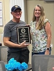 Two people smiling, one holding an award plaque, in a room with a blue decoration on a table.