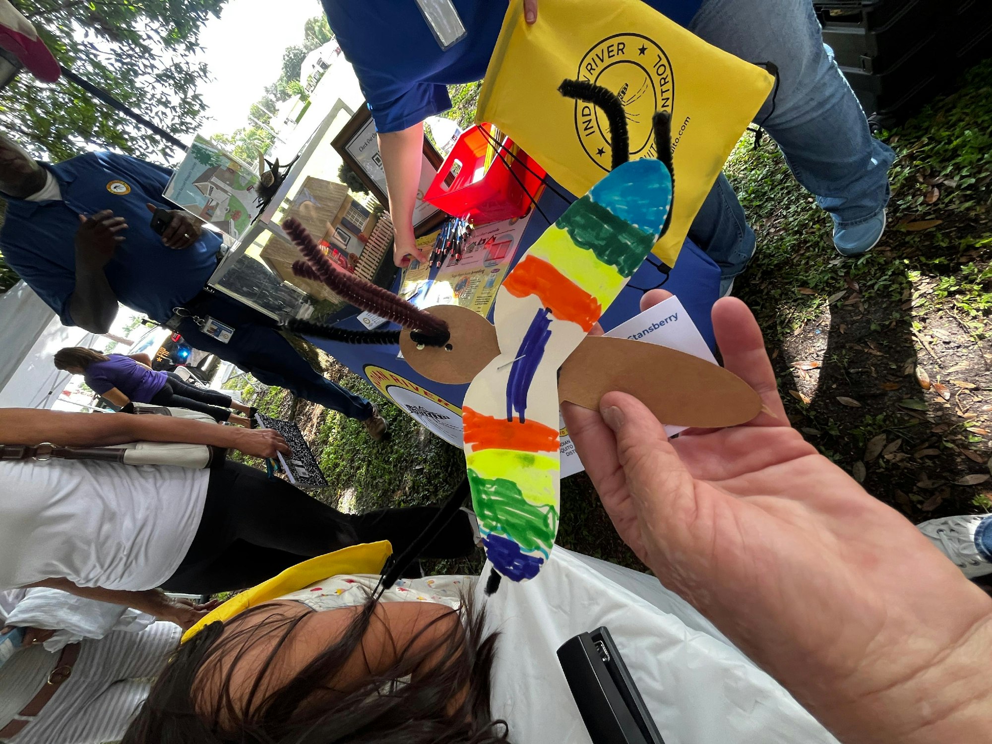 Colorful paper rocket craft held by a hand at an outdoor event with people.