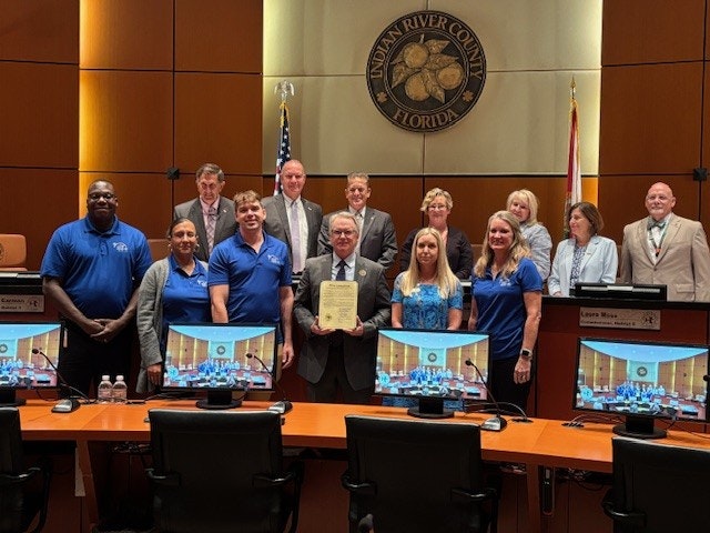 A group of people in a conference room with a seal of Indian River County, Florida, displayed behind them. One holds a certificate.