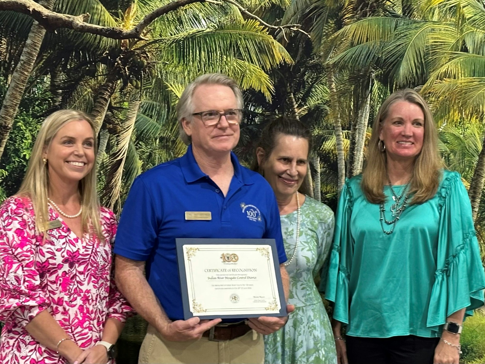 Four people standing together, one holding a certificate, with a palm tree backdrop.