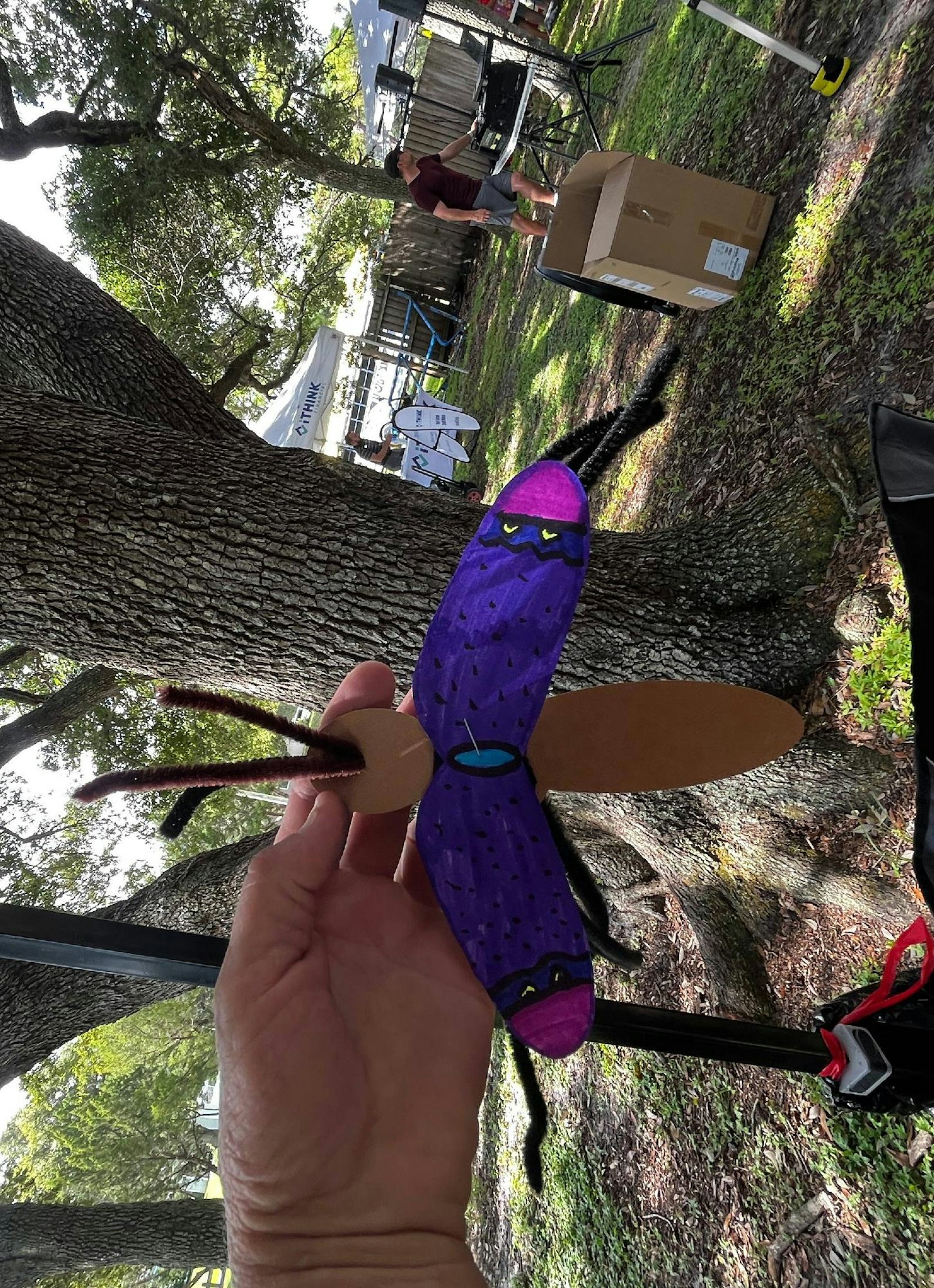 A hand holding a creative, colorful craft resembling a character with an outdoor setting in the background.