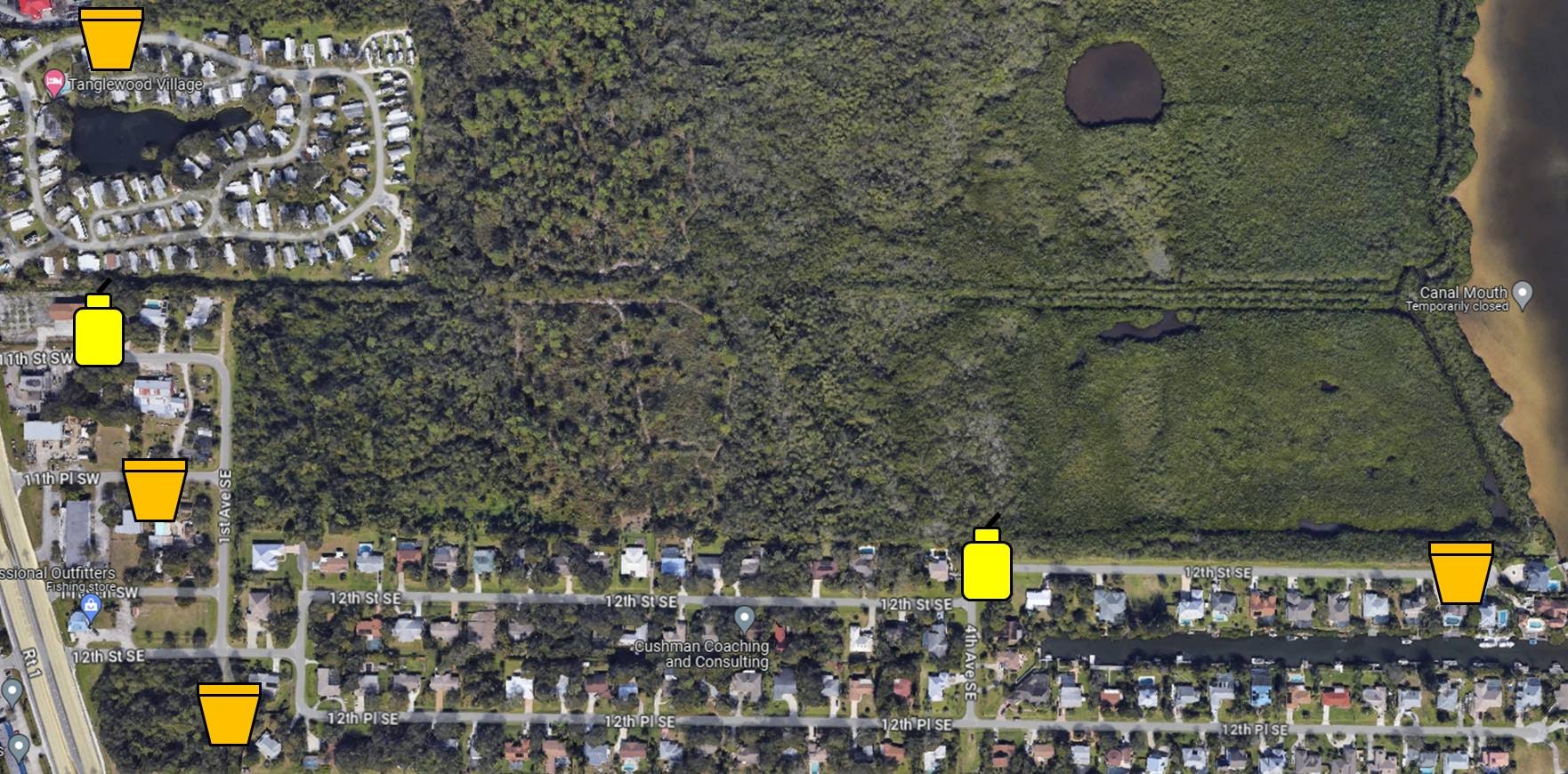 Aerial view of a residential area with roads, houses, wooded land, and small bodies of water. Yellow icons probably indicate specific points of interest.