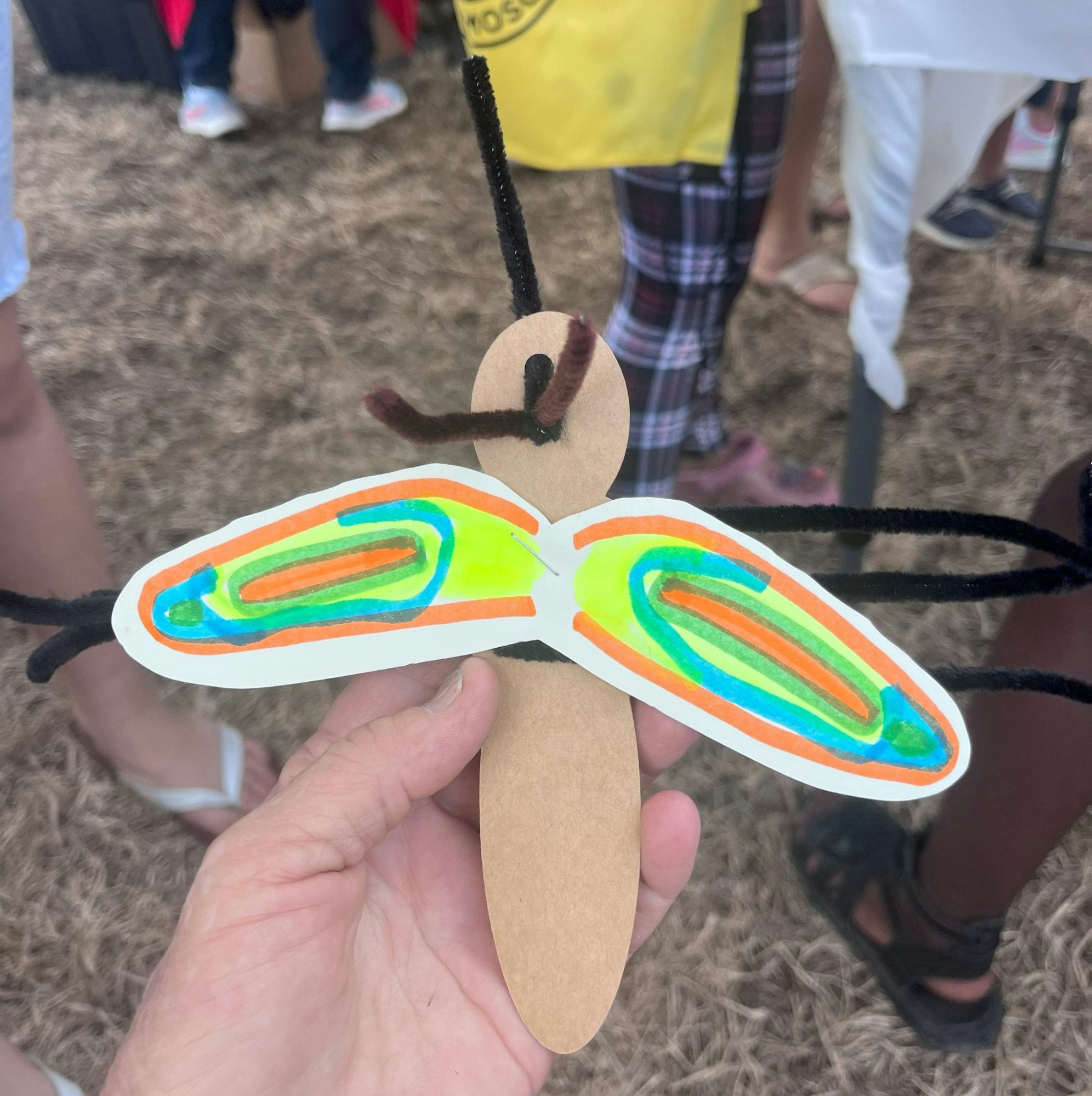 A hand-held, craft-made paper butterfly with colorful wings and pipe cleaner antennae, with people's legs in the background.