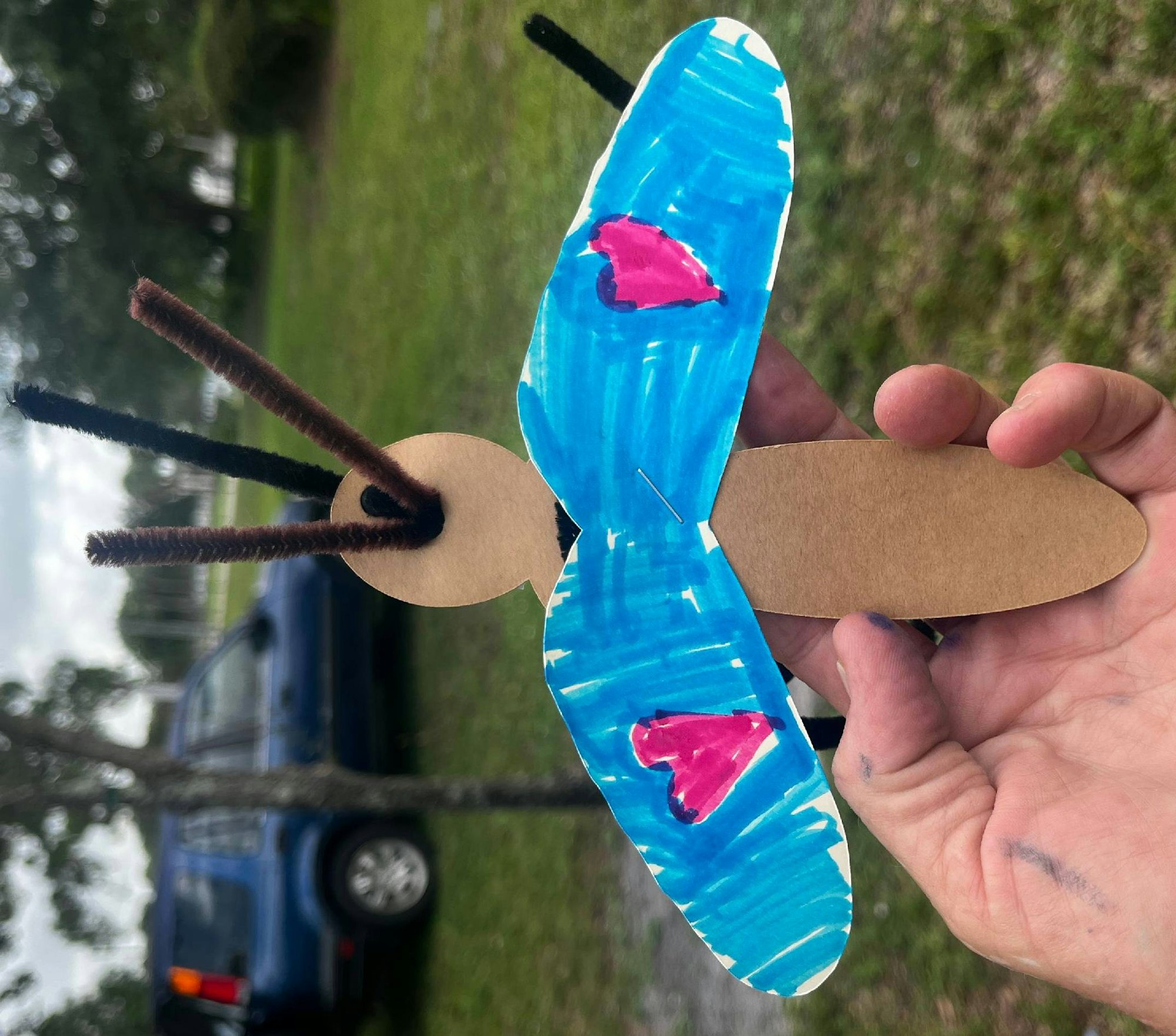 Handmade butterfly craft with cardboard body, pipe cleaner antennae, and painted wings held by a hand.