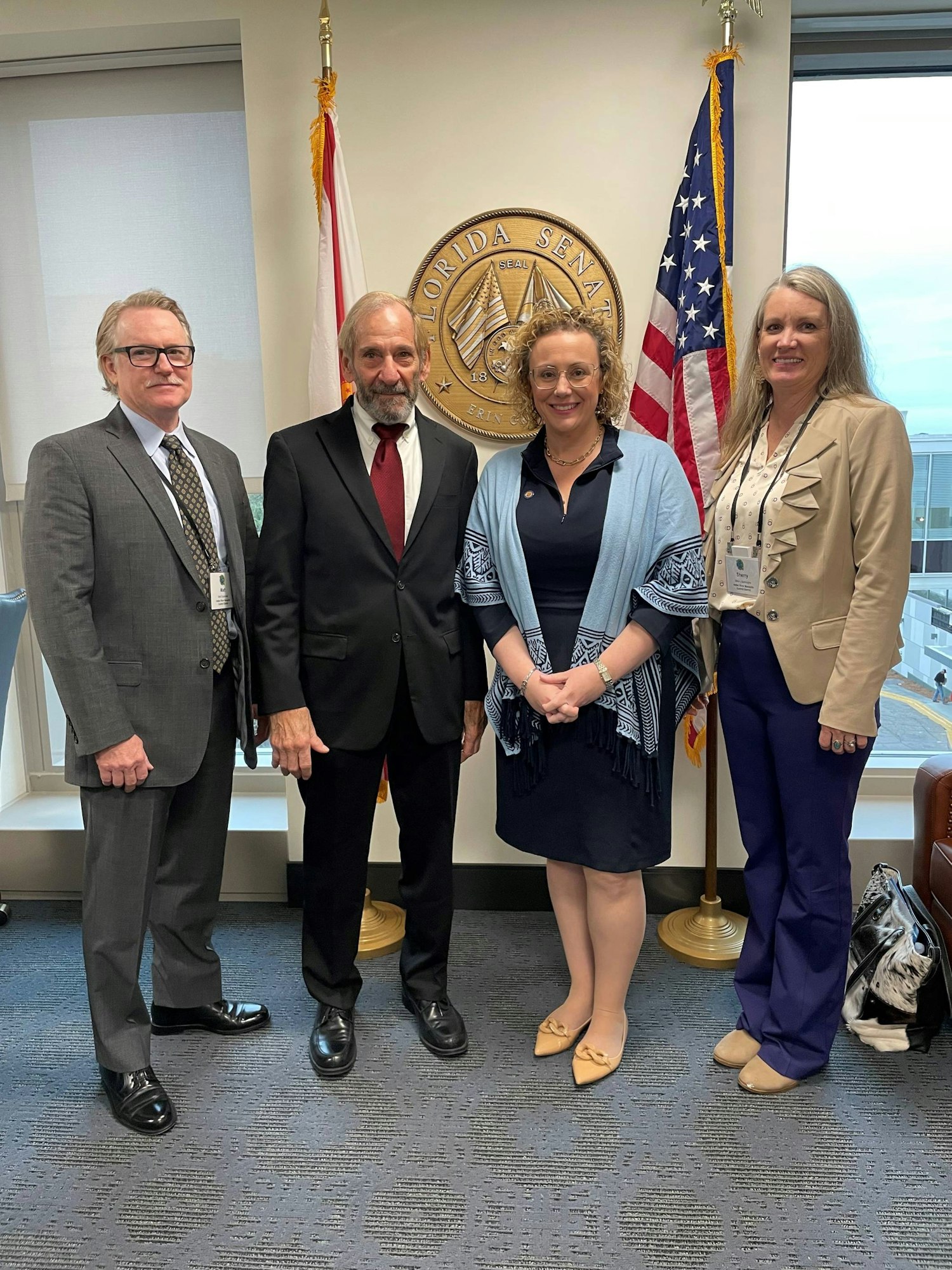 Four individuals posing in an office with the Florida Senate seal and US and state flags behind them.