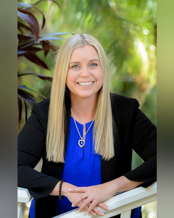 A smiling woman with long blonde hair, wearing a blue top, black jacket, and a heart-shaped necklace with a greenery backdrop.