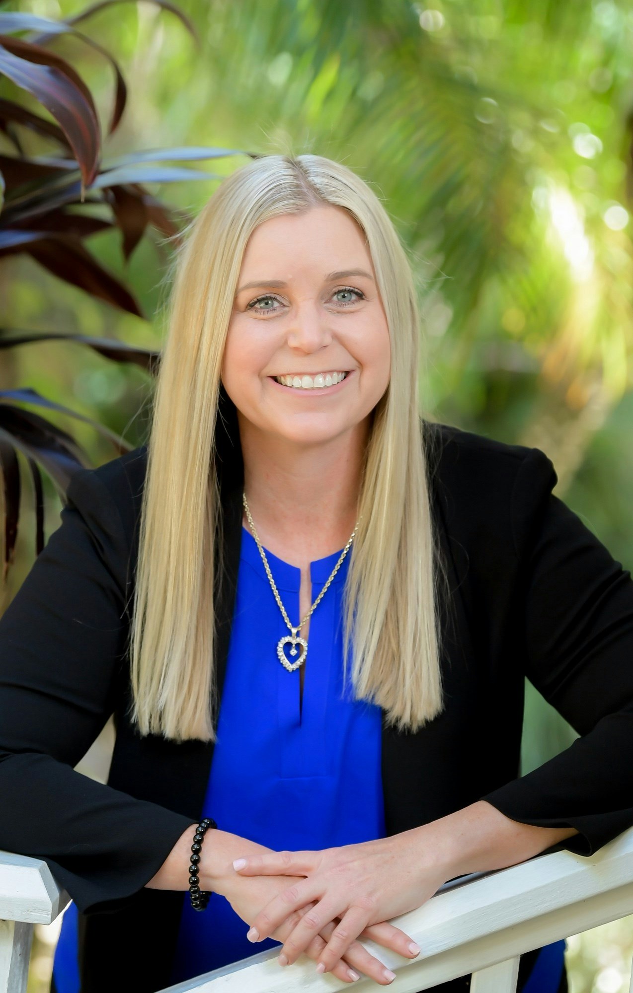 A smiling woman with long blonde hair, wearing a blue top, black jacket, and a heart-shaped necklace with a greenery backdrop.