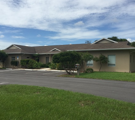 A single-story building with a parking lot, green lawn, blue sky, and clouds.
