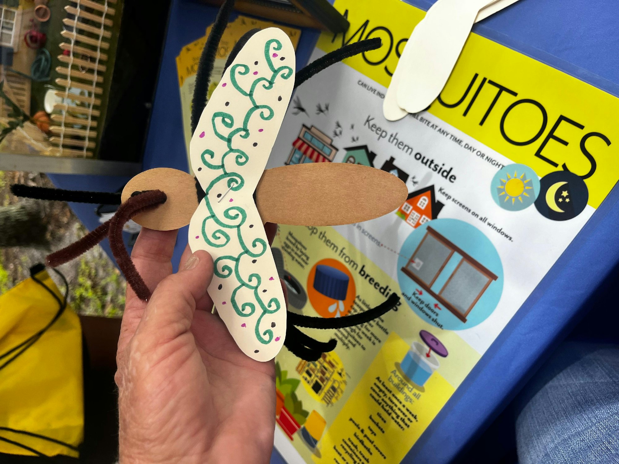 A hand holding a craft mosquito against a background with information about mosquitoes.