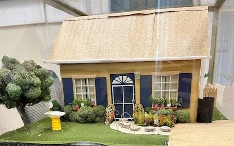 A miniature house model with a beige exterior, blue trim, front garden, and decorative items like a birdbath and topiaries.
