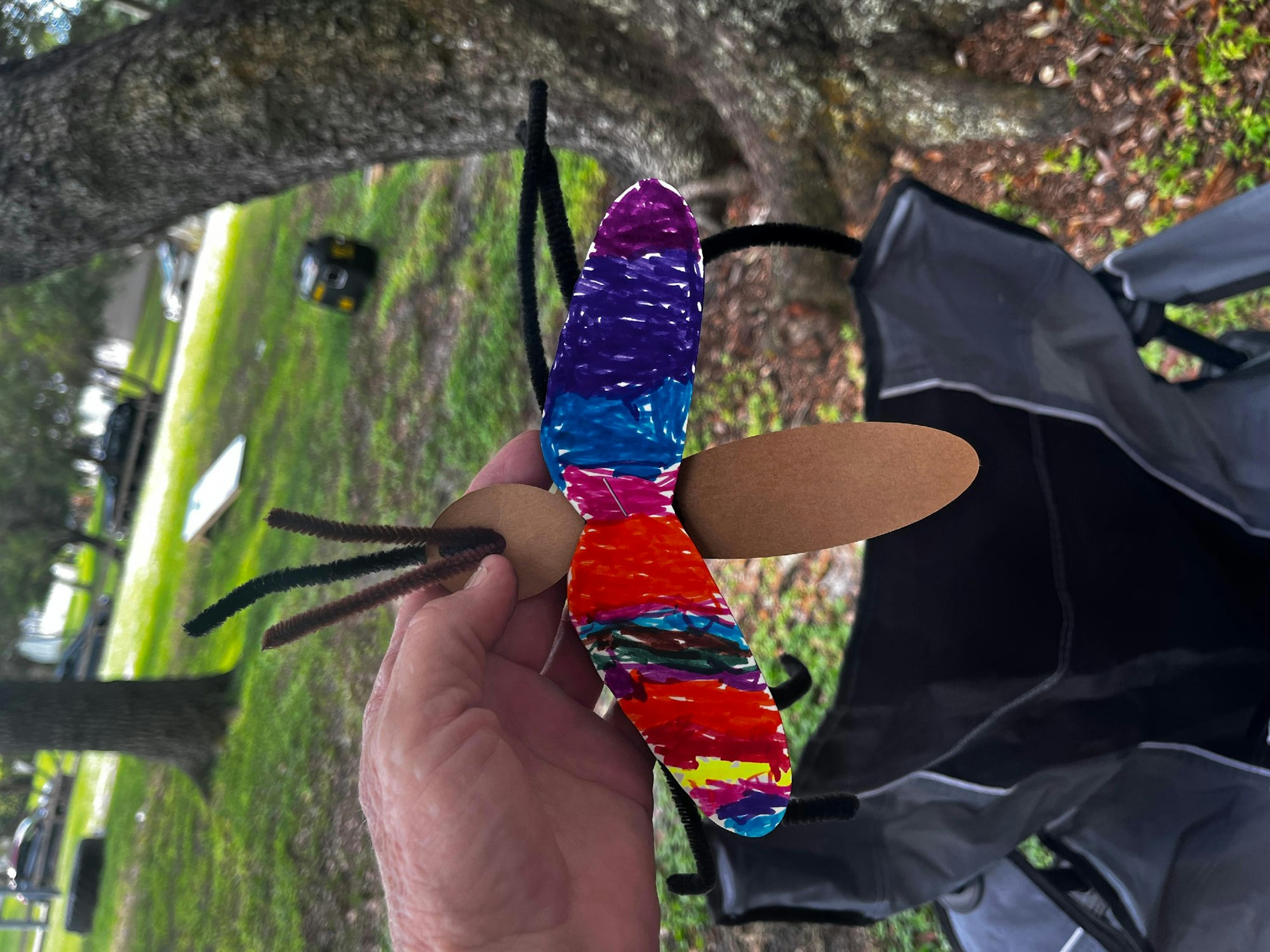 A colorful handmade butterfly with pipe cleaner antennae, held by a person outdoors.