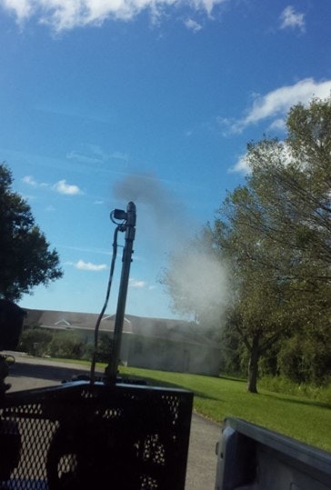 A pipe emitting steam or smoke outdoors near trees under a clear blue sky.