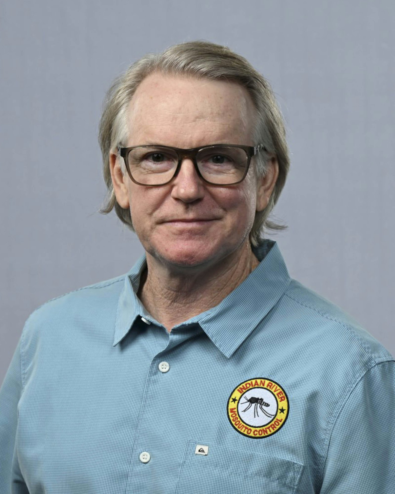 A man in a blue shirt with "Indian River Mosquito Control" patch, wearing glasses, against a grey background.