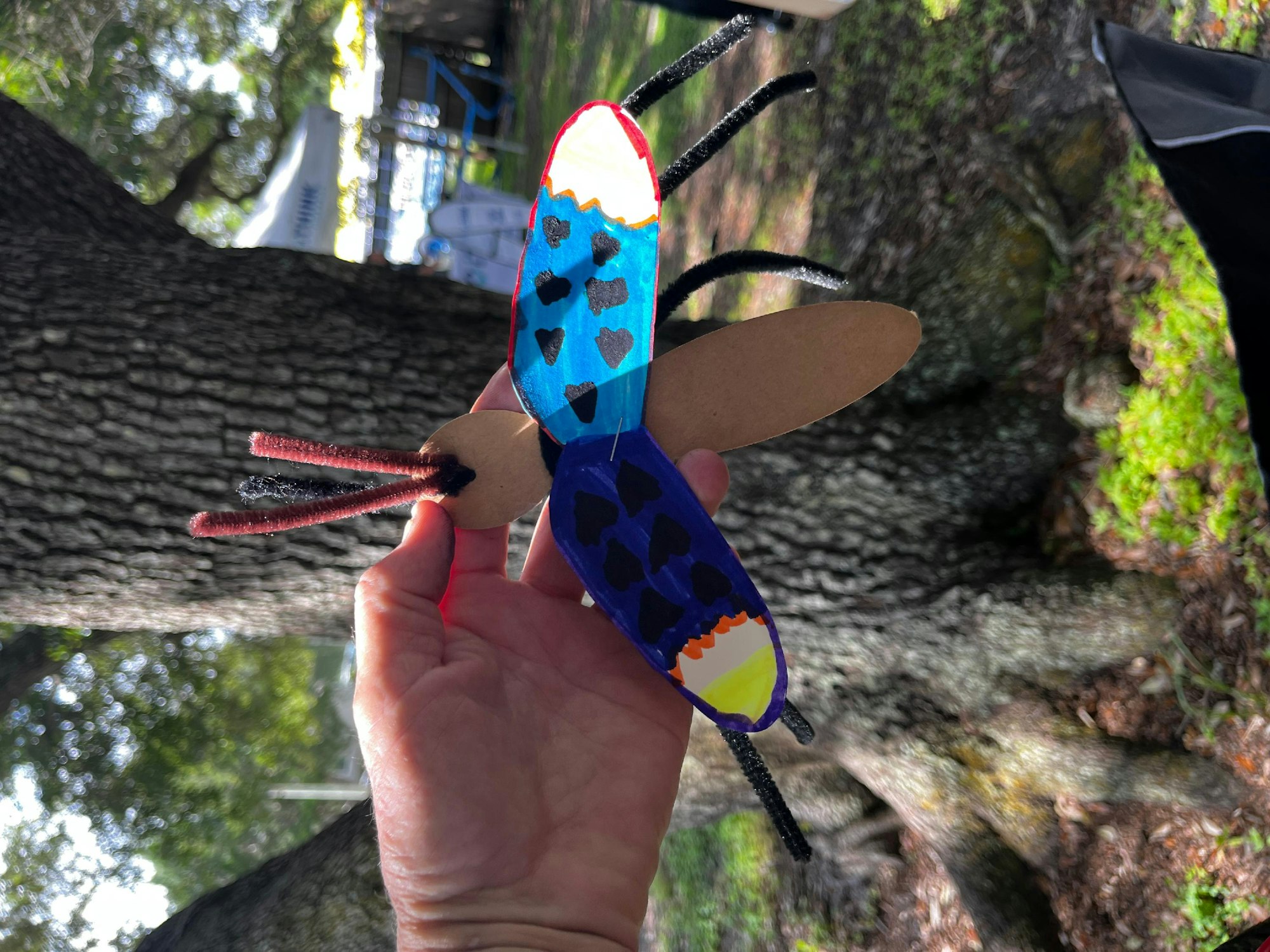 A hand holding a colorful handmade paper butterfly with a nature background.