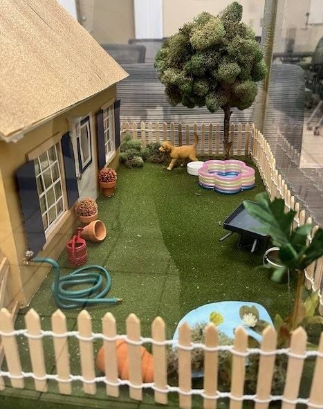 Miniature backyard with a house, tree, plants, fence, hose, and a dog by a pool.