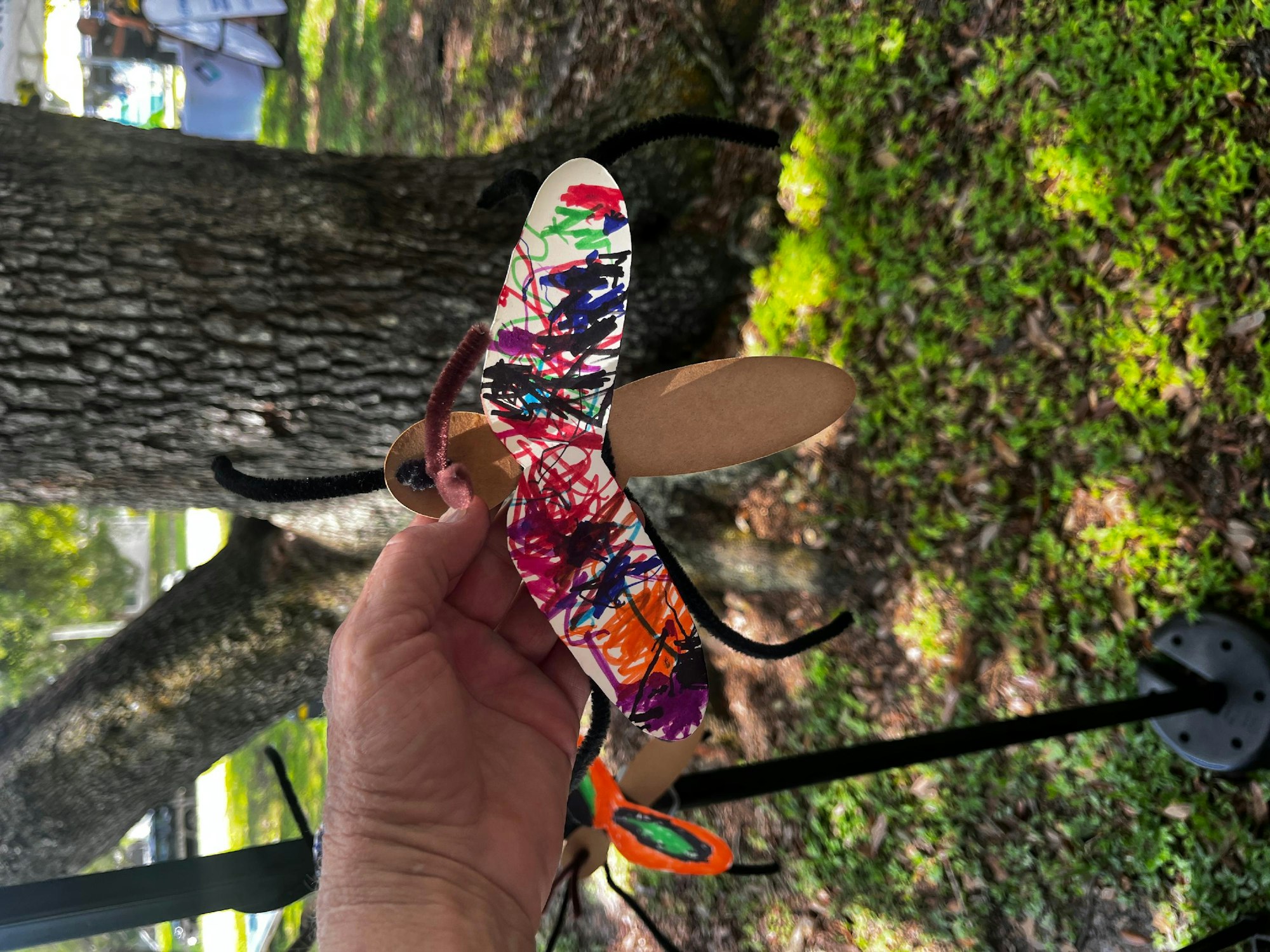 A hand holds a colorful, handcrafted butterfly craft with a tree bark and green moss in the background.